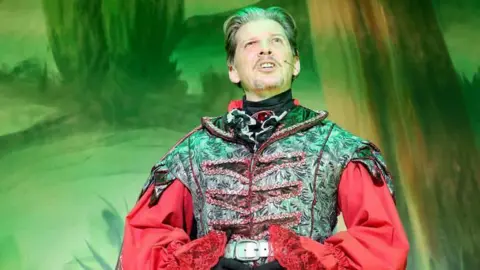 Barry Rivett Hotshots Photography A waist upwards image of Nigel Harman shows him in a black and red Fleshcreep costume. His shiny black coat waistcoat has red braiding and he wears a red shirt underneath with wide sleeves. He has short, dark brown hair swept back from his face with a small moustache and beard and green eyes. He is singing out to the audience.