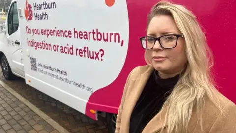 A woman with blonde hair with black glasses in front of a white and pink van which reads: DO YOU EXPERIENCE HEARTBURN, INDIGESTION OR ACID REFLUX?