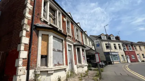 An empty home which has its windows boarded up. The door handle is hanging off. Paint is peeling away. Weeds are growing out of the paved area in front. 