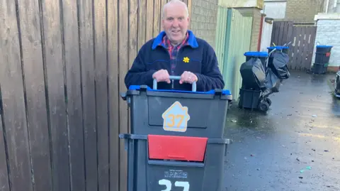 Lawrence Hutchins is holding several box shaped bins that are stackde on top of each other. 