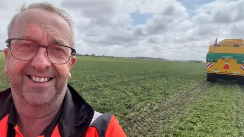 Ian Watson Ian has thinning grey hair and is standing in a pea field wearing an orange high-vis jacket. He is on the far left of the shot. On the far right is the rear of a yellow combine harvester