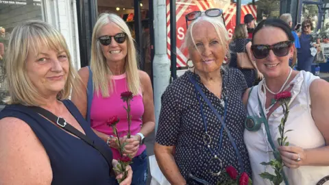 Four woman stood on the side of the road, waiting to pay their respects to Bonnie Tyler. They are each holding pink flowers. They are all smiling and looking at the camera.