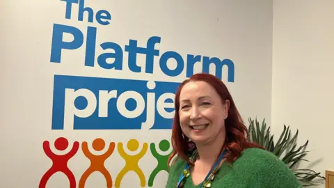 Image shows Sadie Sharp, CEO of The Platform Project. Her hair is shoulder length, curled and red in colour. She is to the right of the photo smiling. She has a green V-neck knitted jumper on. Behind her is a green plant. On the white wall behind her, there is a large 'The Platform Project' sticker. The words are blue. Underneath, there are stick figure stickers in red, orange, yellow and green. 