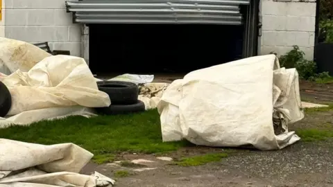 Himley Cricket Club Tyres and sheeting are scattered across grass and gravel in front of an outbuilding with a damaged door.