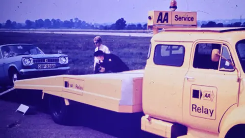 The AA An old colour photo of a yellow AA Relay lorry with a blue car being put onto the bed with a patrolman in uniform standing next to the truck beside the motorist