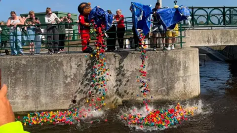 A rainbow stream of different coloured rubber ducks are being poured into the River Tees by organisers - one who is wearing a bear costume. Onlookers lean over metal railings and film with their phones.