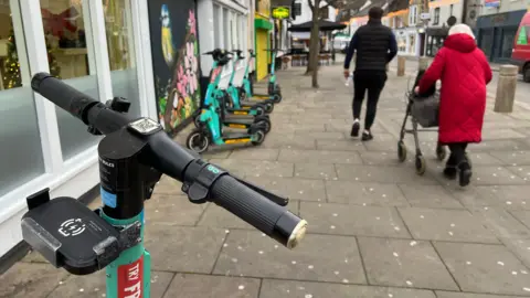 Simon Dedman/BBC E-scooters parked on a wide pavement in a city centre with an old lady in large road coat walking past them aided with a walker.