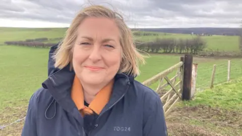 Alison Hume smiles at the camera, with some green fields behind her and blackened moorland in the distance.