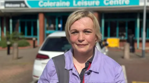 A blonde woman, with a lilac jacket stares at the camera with a neutral expression