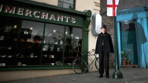 A police officer in Victorian costume standing in a street with a lamp post beside him and a bicycle leaning on the wall of a Victorian-style shop with large windows
