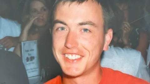 Martin as a young man, he is in a crowded room and is wearing an orange tshirt.