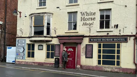 A three storey building in poor condition. The cream exterior is cracked and the windows on the first floor are smashed and the woodwork rotten. A man can be seen standing outside a fake red front door in the middle of the building. Two signs read: "White Horse Hotel".