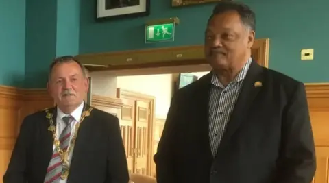 El reverendo Jesse Jackson viste traje negro y camisa blanca a rayas. Tiene cabello corto y negro y bigote. Maolíosa McHugh también aparece en la foto con traje negro y corbata a rayas rojas y grises. Lleva sus cadenas doradas de alcalde.  
