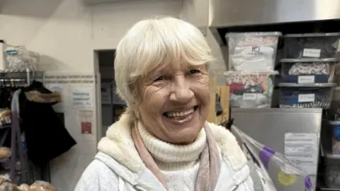 A lady smiling with light blonder hair wearing a grey furry zip up hoody and a pink and grey scarf. 