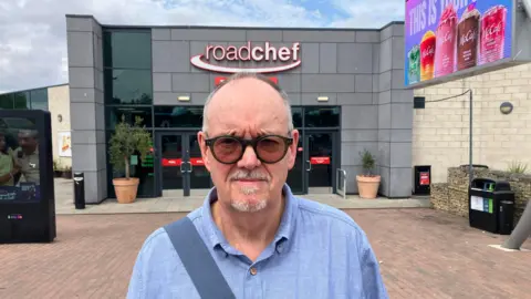 A man wearing a lilac shirt and sunglasses stands outside a square building with a Roadchef logo. 