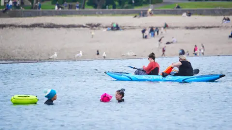 PA Two woman sit in a kayak while two others swim in Clevedon Lake, while people sit and stand on the beach 