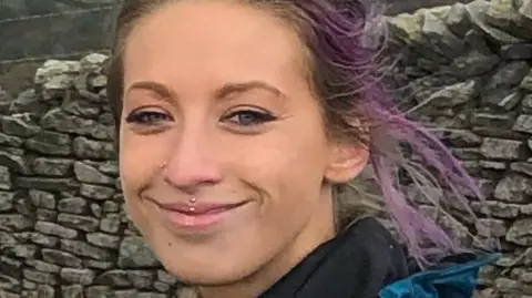 Kristie Hunt, who has brown hair with purple highlights and a piercing in her top lip, smiles at the camera in a countryside setting with a stone wall in the backdrop.