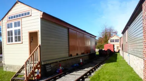 Christopher Hill This picture has a green and brown shed and a railway track in vision. The shed has multiple windows The shed has wooden steps leading to the front door. Other green outbuildings surround the shed. 