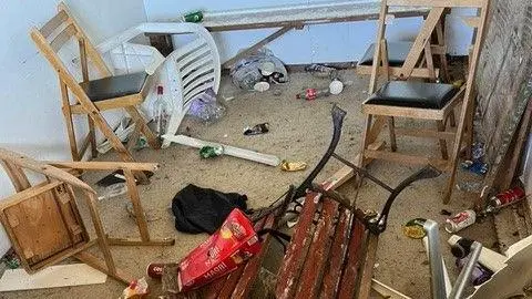 Tarbarrow Cricket Club A disordered room with debris, featuring two wooden chairs with black seats, a broken bench with red slats and a black metal frame, an overturned white plastic chair, and an office chair. The floor is littered with rubbish including bottles, cans, food wrappers, and a broken piece of plasterboard.