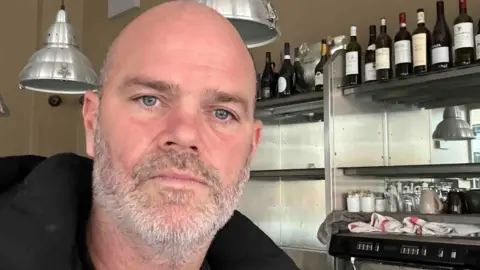 Joel is a bald man with a short grey beard and blue eyes. The selfie is taken in front of metal shelving with bottles of wine on them.