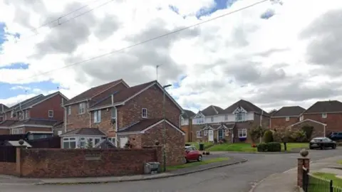 Google A row of brown-bricked semi-detached houses behind a brick fence in the village.