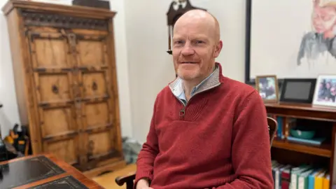 BBC Gavin St Pier looks at the camera he is wearing a red jumper with a quarter zip. He is bald and has a very short and neat ginger beard.