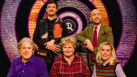 Alan Davies, Phil Wang, Sandi Toksvig, Tom Allen and Sara Pascoe pose in front of the QI logo