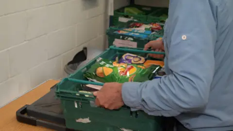 Cambridge City Foodbank A person wearing a long sleeve blue shirt is holding a green tray which has been filled with food. They are lifting it off a wooden table. At the top is a green packet of crisps and some white chocolate buttons in a purple packet.