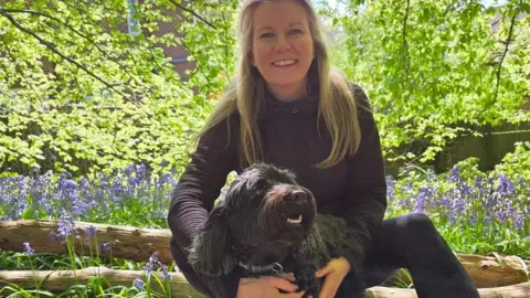 A woman with blonde hair sitting outdoors in a wooded garden area, holding a black dog in front of her. She is smiling and wearing dark athletic-style clothing.