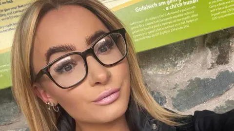 Facebook Close up selfie of Ms Ashton smiling. She has straight blonde hair, black glasses and is wearing a black jacket. She is standing against a stone wall with a green sign behind her. 
