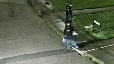 Avon and Somerset Police A man dressed in dark clothing stepping out from a pavement onto the road. He appears to be holding a phone in one hand and a bag in the other. The quality is low and his facial features cannot be made out.