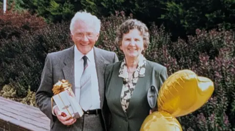 Handout A family photograph showing David wearing a suit, along with his late wife, May, who is holding two golden coloured balloons shaped like a love heart. In the background, there are different coloured flowers in bloom. 