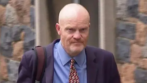 BBC A man with a bald head walking towards the camera. He is wearing a navy blazer, blue buttoned shirt and a burgundy patterned tie. 