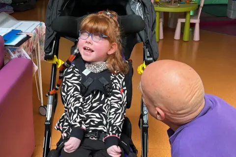 A young girl with ginger hair sits in a pram and grins. She has blue glasses and a Zebra print top on. A red, black and white snake is curling around her shoulders.