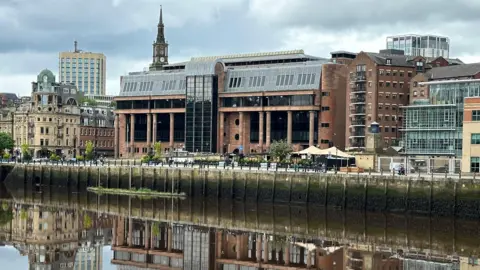Newcastle Crown Court reflected in the River Tyne running in front of it. It is an imposing building made from smooth red stone with massive black windows and tall columns along its frontage.