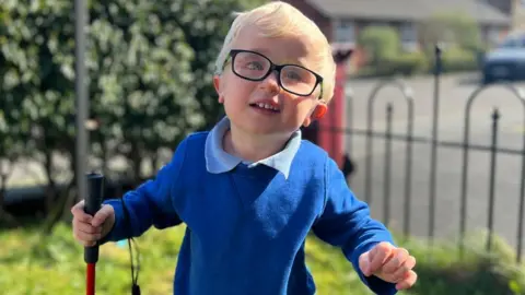 Harvey Hind with blonde hair wearing black glasses and royal blue school sweatshirt over a pale blue polo shirt. He is in the garden in the sunshine and holding a red-and-white striped cane