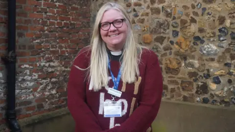 Diocese of St Edmundsbury and Ipswich Samantha Brazier-Gibbs has long blonde hair and is wearing black rimmed glasses, a clerical collar and a burgundy jumper with letters, which are obscured by her lanyard. She is smiling and standing in front of flint and brick walls. 