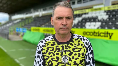 Clab Arrestman Dale Vance dressed in the leopard prints, which contains clean hands. Stands on a football stadium, with the stomach lines behind him. They have dark black hair and looking at the camera and eyes-visible eyes with empty words. 