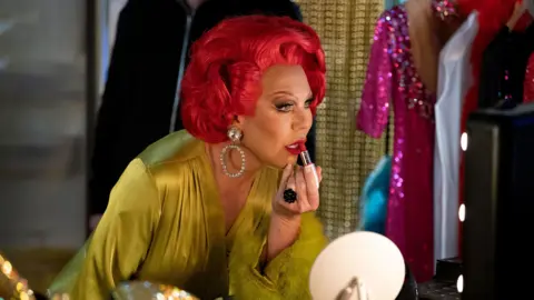 La Voix is applying lipstick to her face in a dressing room