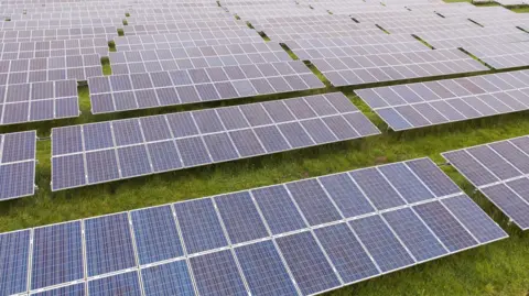 An aerial view of a solar farm with several rows of black panels on top of the grass facing the sunlight.