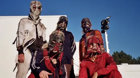 Sam Locke A group of five actors in character as the 'Jimmy' cult members. All ears hoofed masks, with long blonde wigs visible. They all wear ragged tracksuits.