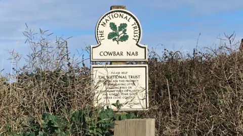 Stuart Arnold/LDRS Cowbar, near Staithes, is adjacent to the Cleveland Way and a popular spot particularly with walkers. A National Trust sign stands in shrubbery with green lettering and oak leaves. The sign reads 'COWBAR NAB'.
