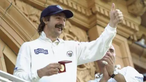 Getty Images Jack Russell - a cricket player who is wearing a long-sleeved cricket top with badges on it and a navy cap with a crest on it - smiles as he holds a medal in a red box with one hand and gives a thumbs up with the other. He has a brown mullet and a moustache. There is another cricket player next to him, clapping. They are stood on a stand in front of an ornate building.