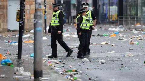 PA Media Police Scotland officers walk along a city street covered in litter and debris following large crowds gathering in Glasgow.