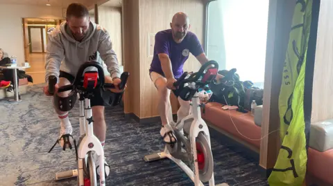 JODIE CANNELL Darren, a man with short brown hair and a grey jumper and Adrian a bald man with a purple t-shirt on exercise bikes on the Manxmman.
