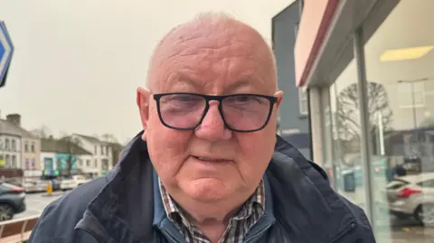 Mr Hawe is a bald headed man who is wearing large black square glasses. He is wearing a navy coat, checked shirt and zip up fleece. The picture is taken on a street with buildings in the background.
