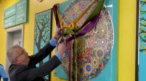 Charles Heslett/BBC A man wearing a suit using scissors to cut some gold, green and purple ribbons hanging in front of circular, multicoloured mosaic.