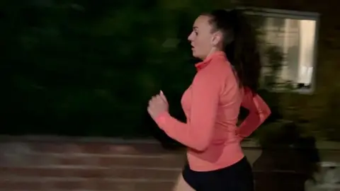 BBC Natalie Bell runs past a house in the dark. She is wearing a pink running top and black shorts.