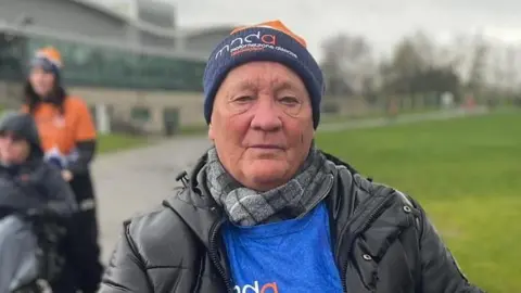 Simon Thake/BBC A man in a navy MNDA hat and blue MNDA shirt. He has a neutral expression. He has a chequered black and white scarf around his neck and a black coat on.