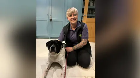 Birmingham Dogs Home A woman with short blonde hair kneels next to a large black and white dog that is lying down. 